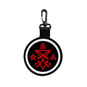 May include: A round black keychain charm with a white border and a red symbol design. The charm has a black strap and a metal clasp. The design includes a pentagram, a triangle, and other geometric shapes.