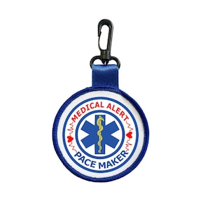 May include: A round medical alert patch with the words "MEDICAL ALERT" and "PACE MAKER" in a blue and white design. The center features a blue Star of Life symbol with a yellow staff. A blue strap and black clip are attached.