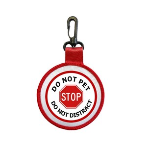Op de afbeelding: Een ronde, rood-witte patch met de woorden "DO NOT PET" en "DO NOT DISTRACT" rond een rood stopbord. De patch is bevestigd aan een rode band en een zwarte clip.
