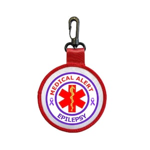 Puede incluir: Un parche redondo de alerta médica con borde rojo. Presenta un símbolo rojo de la Estrella de la Vida y el texto "MEDICAL ALERT EPILEPSY". Incluye una correa roja y un clip negro.