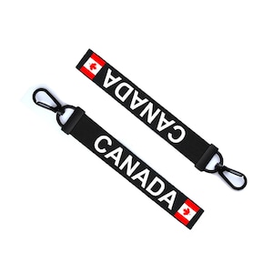 Op de afbeelding: Twee zwarte sleutelhangers met een witte "CANADA" tekst en een Canadese vlag. Elke sleutelhanger heeft een zwarte metalen clip.