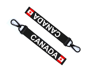 Könnte beinhalten: Zwei schwarze Schlüsselanhänger mit weißem "CANADA"-Text und einer kanadischen Flagge. Jeder Schlüsselanhänger hat einen schwarzen Metallclip.