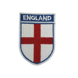 Puede incluir: Un parche bordado con fondo blanco y una cruz roja. El parche tiene forma de escudo y lleva la palabra "ENGLAND" en letras azules en la parte superior.
