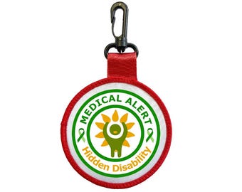 Puede incluir: Un parche de alerta médica redondo, rojo y blanco, con las palabras "MEDICAL ALERT" y "Hidden Disability" en texto verde. El parche presenta una cinta verde y un gráfico amarillo y verde. Tiene un clip negro y una correa roja.