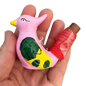 Puede incluir: Un silbato de cer&aacute;mica rosa en forma de p&aacute;jaro con detalles pintados en verde y amarillo. El silbato tiene una cuerda roja atada a una pieza de madera marr&oacute;n.