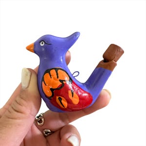 Puede incluir: Un silbato de cer&aacute;mica de color morado en forma de p&aacute;jaro con detalles pintados en naranja y rojo. El silbato tiene una boquilla de arcilla marr&oacute;n.
