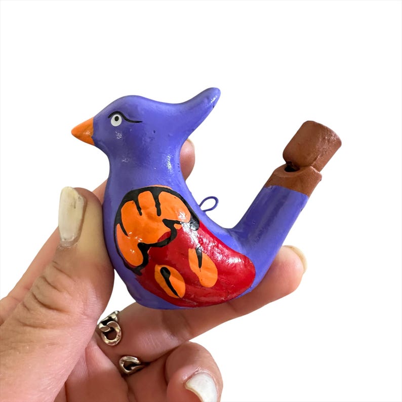 Puede incluir: Un silbato de cer&aacute;mica de color morado en forma de p&aacute;jaro con detalles pintados en naranja y rojo. El silbato tiene una boquilla de arcilla marr&oacute;n.