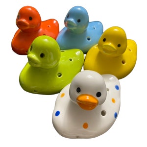 May include: Five ceramic duck figurines in different colors. The ducks are all in a sitting position with their beaks open. The ducks are painted in various colors, including orange, blue, yellow, green, and white. The white duck has blue and orange polka dots.