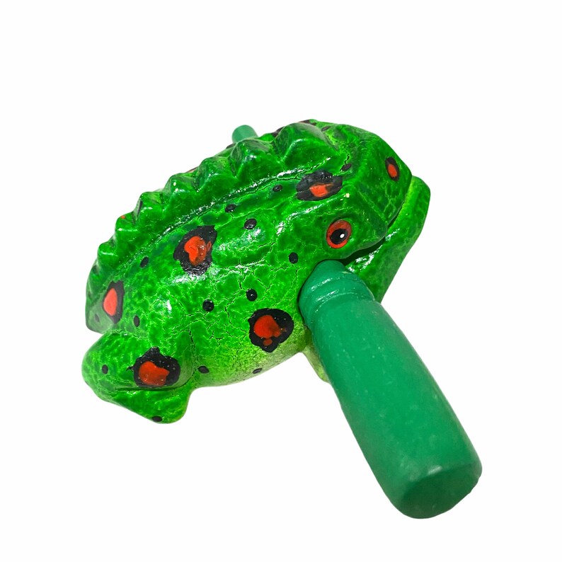 Peut inclure: Grenouille en bois verte avec des taches rouges et noires, tenant un b&acirc;ton en bois vert. La grenouille est un instrument de musique.