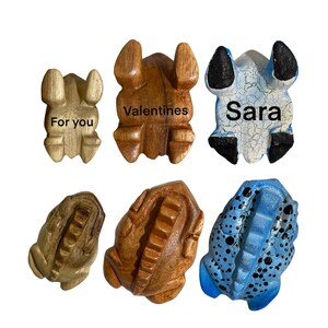 Peut inclure: Six figurines de grenouilles en bois, chacune avec un design et une inscription diff&eacute;rents. Une grenouille est peinte en bleu avec des points noirs, une autre est peinte en bleu et blanc, et les quatre restantes sont en bois naturel. Les inscriptions indiquent "Pour toi", "Saint-Valentin", "Sara", et deux autres qui ne sont pas visibles.