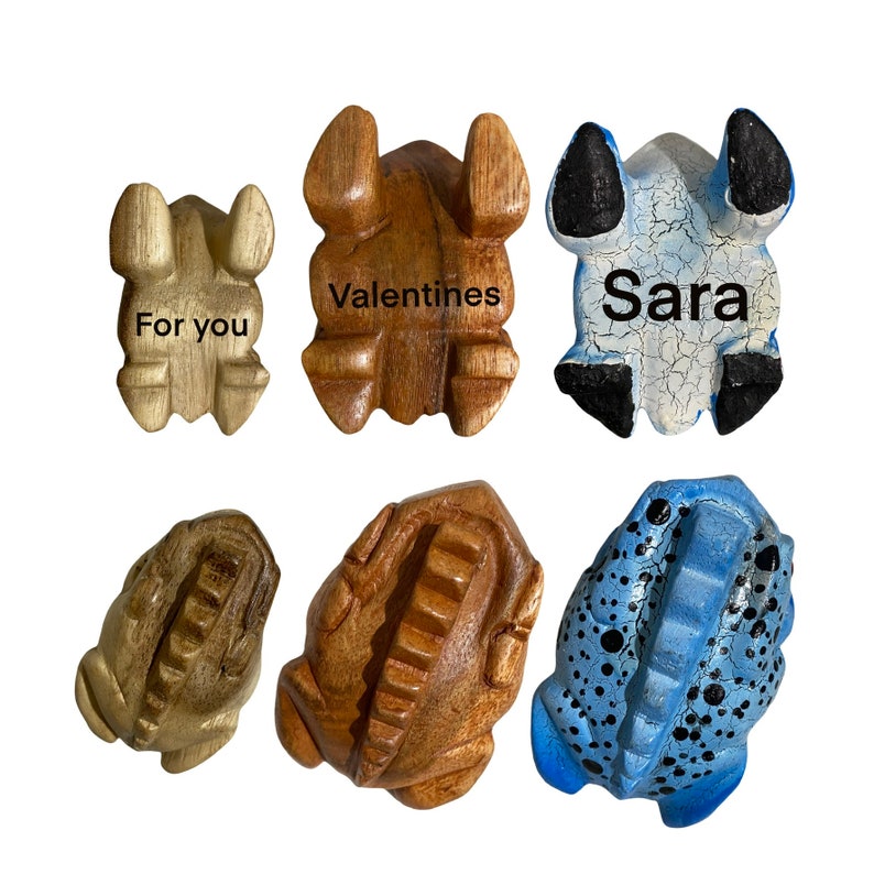 Peut inclure: Six figurines de grenouilles en bois, chacune avec un design et une inscription diff&eacute;rents. Une grenouille est peinte en bleu avec des points noirs, une autre est peinte en bleu et blanc, et les quatre restantes sont en bois naturel. Les inscriptions indiquent "Pour toi", "Saint-Valentin", "Sara", et deux autres qui ne sont pas visibles.