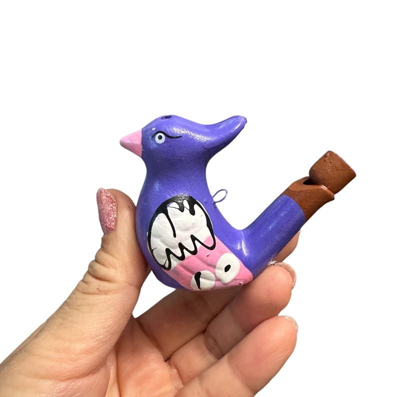 Puede incluir: Silbato de cer&aacute;mica de color morado en forma de p&aacute;jaro con detalles pintados en rosa y blanco. El silbato tiene una boquilla de arcilla marr&oacute;n.