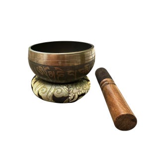 May include: A black and gold singing bowl with a decorative cushion and a wooden striker. The bowl is inscribed with Tibetan characters.