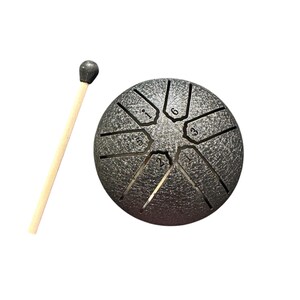 May include: A silver-colored steel tongue drum with numbered cutouts and a wooden mallet. The drum has a textured surface and a rounded shape. The mallet has a dark gray head and a light-colored wooden handle. The drum is designed for musical use.