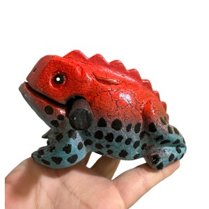 May include: A handcrafted wooden frog figurine. The frog is painted with a red back and a blue belly with black spots. The frog has a textured, cracked appearance and a small, black wooden piece in its mouth.