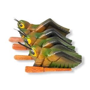 May include: Four wooden grasshopper figurines with brown and green paint and yellow eyes. The grasshoppers are arranged in a row, with their wings spread out. Each grasshopper has a small wooden stick attached to its body.