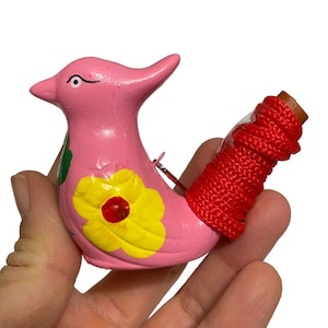 Puede incluir: Un silbato de cer&aacute;mica rosa en forma de p&aacute;jaro con una flor amarilla y una hoja verde pintadas en su cuerpo. El silbato tiene un cord&oacute;n rojo atado a la cola.