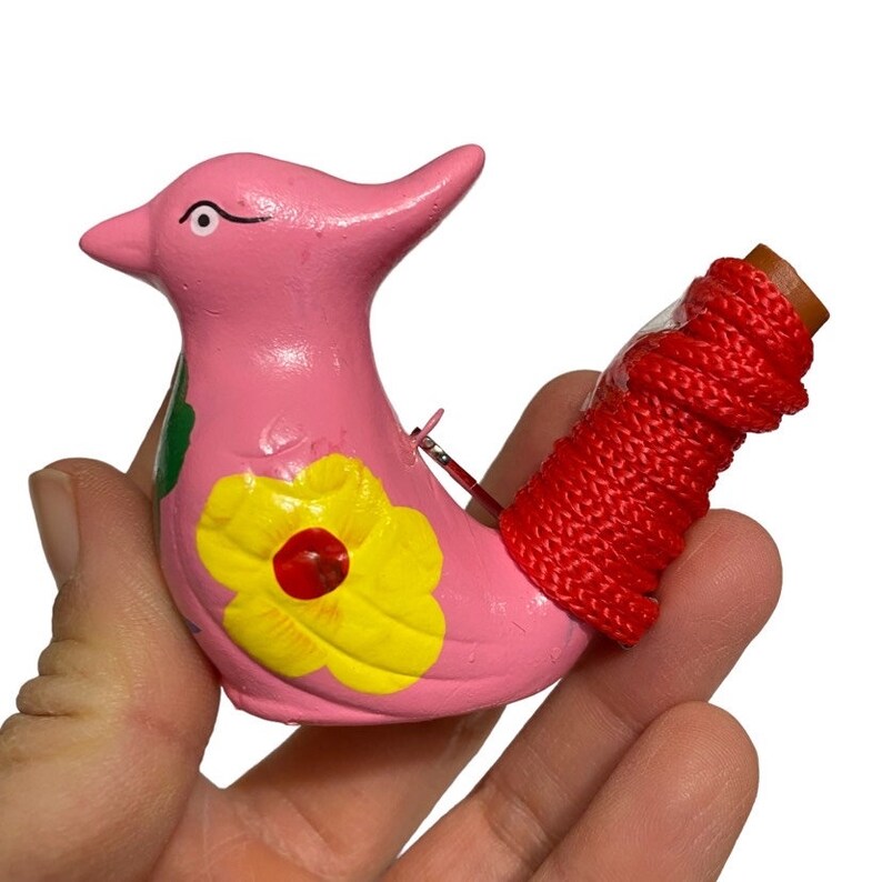 Puede incluir: Un silbato de cer&aacute;mica rosa en forma de p&aacute;jaro con una flor amarilla y una hoja verde pintadas en su cuerpo. El silbato tiene un cord&oacute;n rojo atado a la cola.