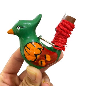 Puede incluir: Un silbato de p&aacute;jaro verde y rojo con detalles pintados de naranja y rojo. El silbato tiene una cuerda roja atada a la parte posterior.