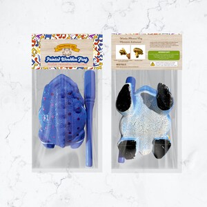 Peut inclure: Un instrument de percussion en bois en forme de grenouille bleue avec un maillet en bois. La grenouille est peinte avec des d&eacute;tails blancs et rouges. L'emballage comprend une &eacute;tiquette qui dit "Cadeau fait main" et "Grenouille en bois peinte".