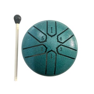 May include: A teal steel tongue drum with a mallet. The drum has a circular shape with eight cut-out tongues and numbers. The mallet has a wooden handle and a dark gray, rounded head. The drum is a musical instrument.