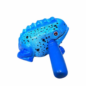 Peut inclure: Un instrument de musique en bois en forme de grenouille bleue avec une surface textur&eacute;e et des taches noires. La grenouille a un &oelig;il rouge et une poign&eacute;e bleue.