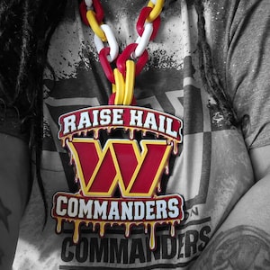 May include: A large pendant featuring the Washington Commanders logo with a dripping effect, inscribed with RAISE HAIL and COMMANDERS, suspended from a chunky plastic chain with alternating red, white, and yellow links over a grey t-shirt.