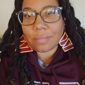 May include: A person wearing large, geometric earrings. The earrings are a deep burgundy colour with gold trim. The person is wearing a burgundy hoodie with a gold and white design. They are also wearing glasses with a dark frame.