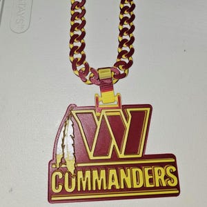 May include: A pendant necklace featuring the Washington Commanders logo in burgundy and yellow. The pendant has the team's "W" logo and the word "Commanders". The chain is burgundy and yellow.