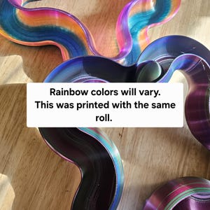 Può includere: Opere d'arte astratte e ondulate, stampate in 3D, in vibranti colori arcobaleno. I pezzi sono disposti su una superficie di legno. Il testo sull'immagine recita: "I colori dell'arcobaleno variano. Questo è stato stampato con lo stesso rotolo."