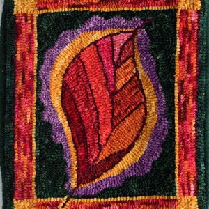 May include: A close-up of a colorful, hand-stitched rug with a single, stylized leaf in the center. The leaf is a vibrant red, orange, and yellow, with a purple outline. The rug is framed with a dark green border and a red, orange, and yellow border.