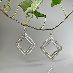 Puede incluir: Un par de pendientes de plata con un diseño de doble cuadrado. Los pendientes están colgados de una rama con hojas verdes.