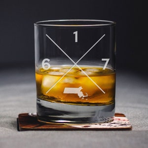 May include: A clear glass with a cross design etched on the side. The glass is filled with amber liquid and ice cubes. The numbers 1, 6, and 7 are etched on the glass. The state of Massachusetts is also etched on the glass.