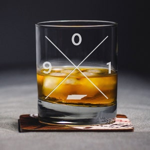 May include: A clear glass tumbler with a white cross design and the numbers 901. The glass is filled with amber liquid and ice cubes. The glass is sitting on a brown coaster.