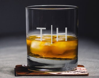 May include: A clear glass with ice and a golden liquid inside. The glass has the letters "T|H" etched on the side. The glass is sitting on a brown coaster.