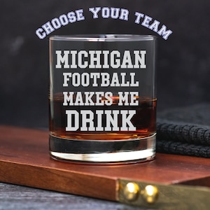 May include: A clear glass tumbler with the text "MICHIGAN FOOTBALL MAKES ME DRINK" in white block letters. The words "CHOOSE YOUR TEAM" are arched above the main text. The glass is filled with a brown liquid, likely a beverage.
