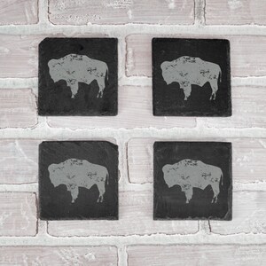 May include: Four square, black slate coasters with a gray bison silhouette design. The coasters are arranged on a white brick background. Each coaster has a unique, natural edge.