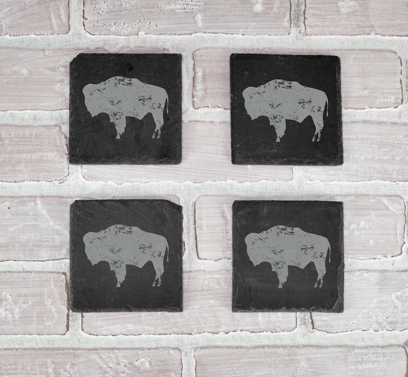 May include: Four square, black slate coasters with a gray bison silhouette design. The coasters are arranged on a white brick background. Each coaster has a unique, natural edge.