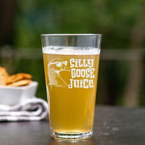 Może przedstawiać: Szklanka do piwa z przezroczystego szkła z białym wzorem gęsi w okularach przeciwsłonecznych i tekstem "Silly Goose Juice". Szklanka jest wypełniona jasnożółtym płynem.