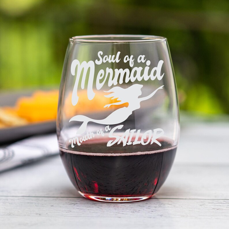 May include: A stemless wine glass filled with red wine. The glass has white text that reads "Soul of a Mermaid, Mouth of a Sailor" with a mermaid silhouette. The glass is on a light-colored wooden surface.