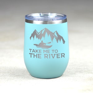 Puede incluir: Un vaso de acero inoxidable turquesa con tapa plateada. El vaso tiene un gr&aacute;fico de una persona en kayak frente a monta&ntilde;as y &aacute;rboles. El texto "Take me to the river" est&aacute; impreso debajo del gr&aacute;fico.
