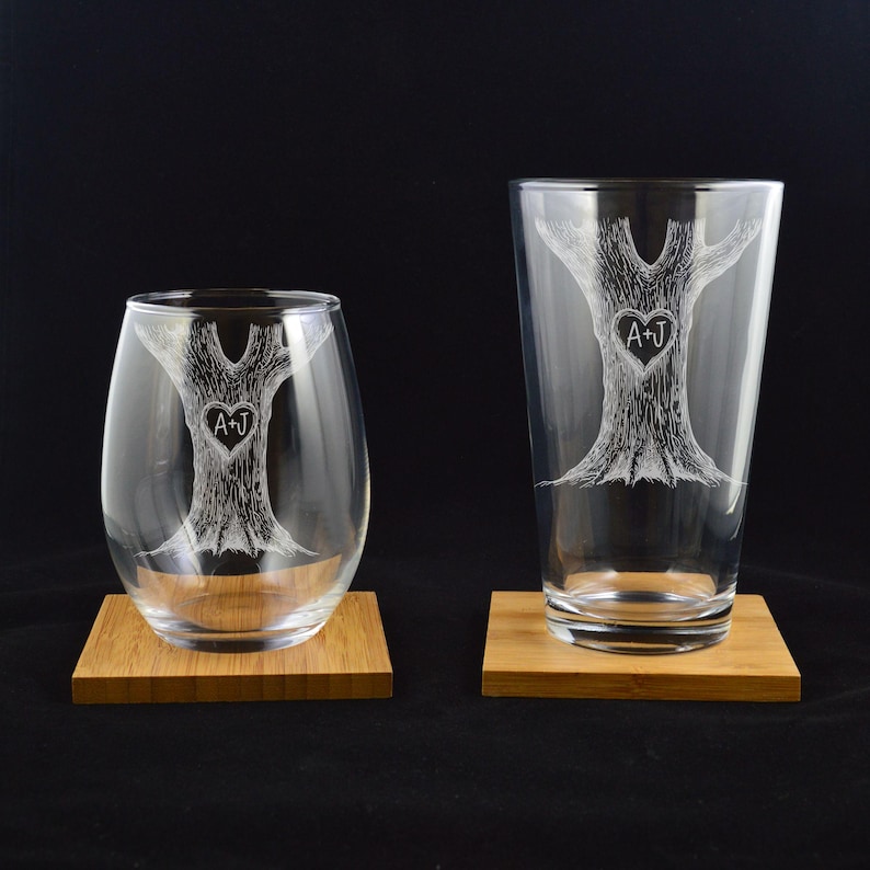 Puede incluir: Dos vasos de vidrio grabados con un dise&ntilde;o de &aacute;rbol y las iniciales A+J en un coraz&oacute;n dentro del &aacute;rbol. Los vasos est&aacute;n sobre posavasos de madera.