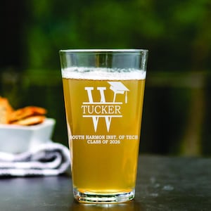 May include: A clear pint glass filled with a light-colored beverage. The glass is etched with the name "Tucker", a graduation cap, and the text "South Harmon Inst. of Tech Class of 2026". The glass is on a dark surface.