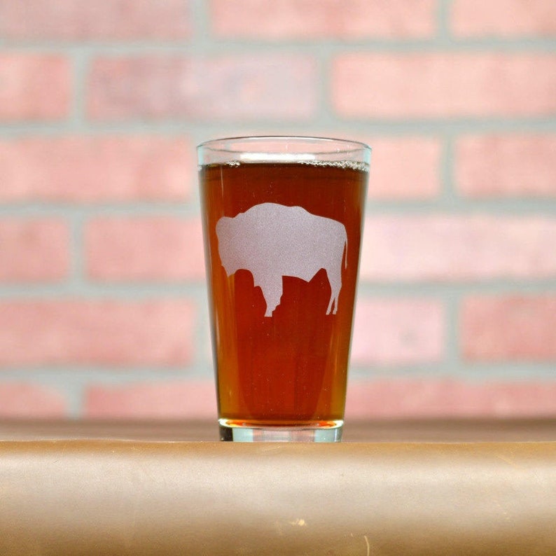 May include: A clear glass pint glass with a frosted silhouette of a buffalo on the side. The glass is filled with a dark amber colored beverage.