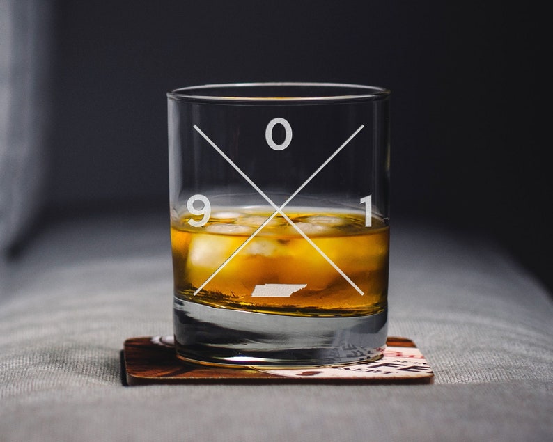 May include: A clear glass tumbler with a white cross design and the numbers 901. The glass is filled with amber liquid and ice cubes. The glass is sitting on a brown coaster.
