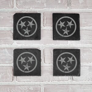 May include: Four black slate coasters with a white circle and three white stars in the center. The stars are arranged in a triangular pattern.