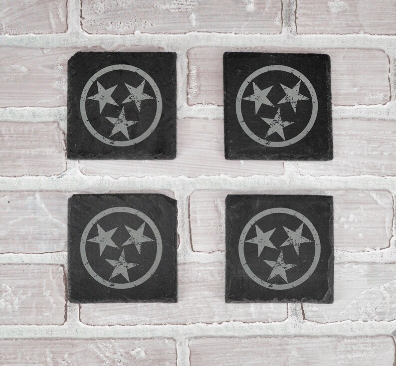 May include: Four black slate coasters with a white circle and three white stars in the center. The stars are arranged in a triangular pattern.