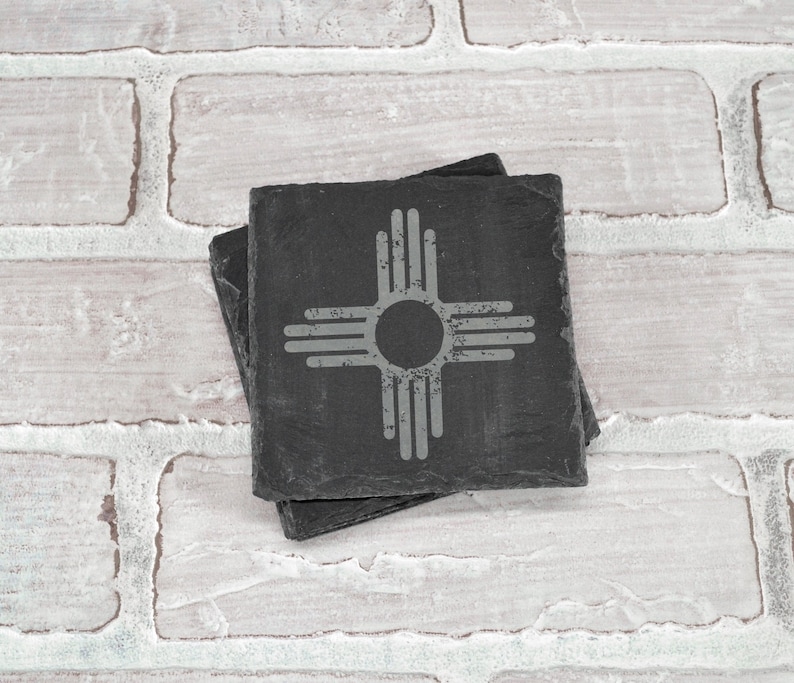 May include: Two black slate coasters with a white Zia symbol, the state symbol of New Mexico, on the top coaster.