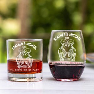 May include: Two clear glasses, one a rocks glass and the other a stemless wine glass, each with a white skeleton couple design. The text "HEATHER & MICHAEL 11.19.11" and "TIL DEATH DO US PART" are printed on the glasses. The rocks glass contains a reddish-brown liquid.