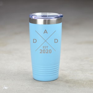 May include: A light blue stainless steel tumbler with a silver lid. The tumbler has an engraved design that says "DAD EST 2020" with a cross and the letters "D A D" at the top.